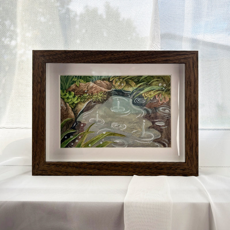 Rainy Day at the Pond — rain landing on a still pond between mossy rocks and reeds